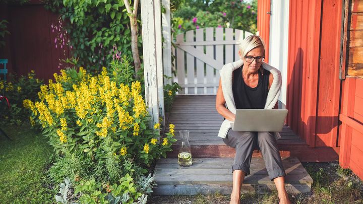 Kvinna sitter vid rött hus med laptop i knät.