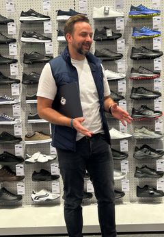 Jag vet inte vem personen är, men bilden visar en man som står framför en vägg med skor. Han har en laptop under ena armen och gestikulerar med den andra handen.