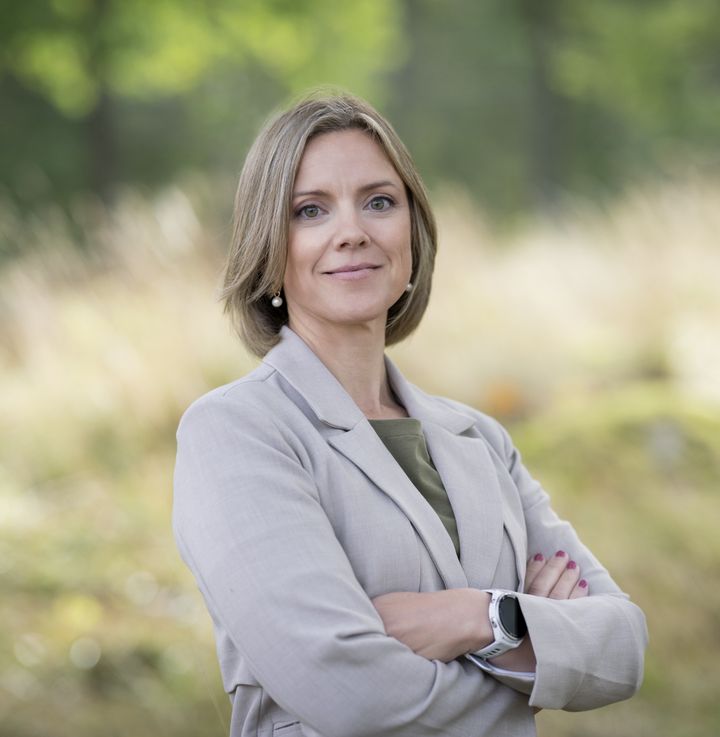 Ann-Louise står utomhus med armarna i kors och ser in i kameran. Bakgrunden är suddig natur med gröna och beige toner.