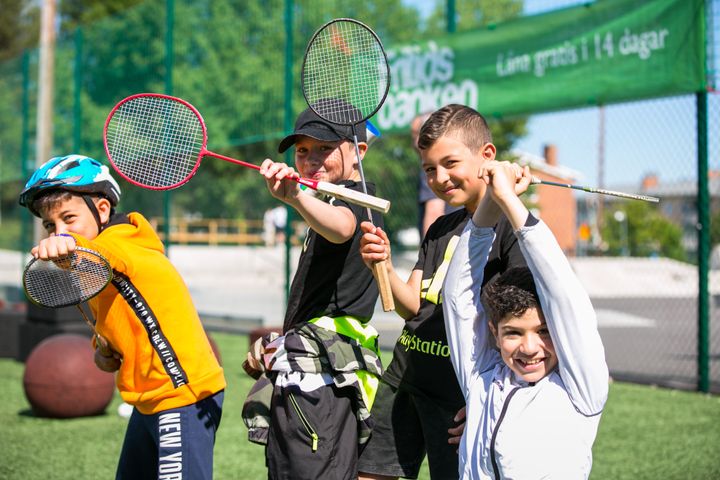 Fyra barn spelar badminton utomhus framför en Fritidsbanken-skylt.