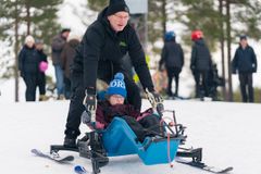 En av Fritidsbankens anställda hjälper ett barn i en sitski på en snöig backe. Flera personer i bakgrunden.