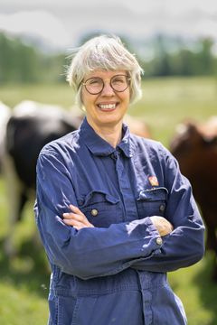 Kvinna i blåställ ute i en kohage, med armarna i kors.