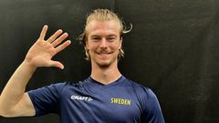 En person med blå tröja som har texten "Sweden" vinkar och ler mot kameran.