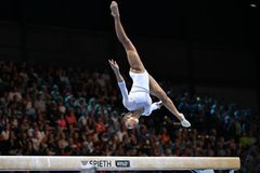 En kvinna i vit dräkt utför en akrobatisk rörelse på bom under en gymnastiktävling, med en stor publik i bakgrunden.