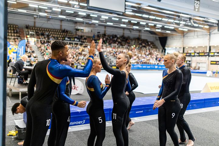 En grupp gymnaster i svarta tävlingsdräkter ger varandra en high-five vid sidan av en gymnastikmatta i en inomhusarena.