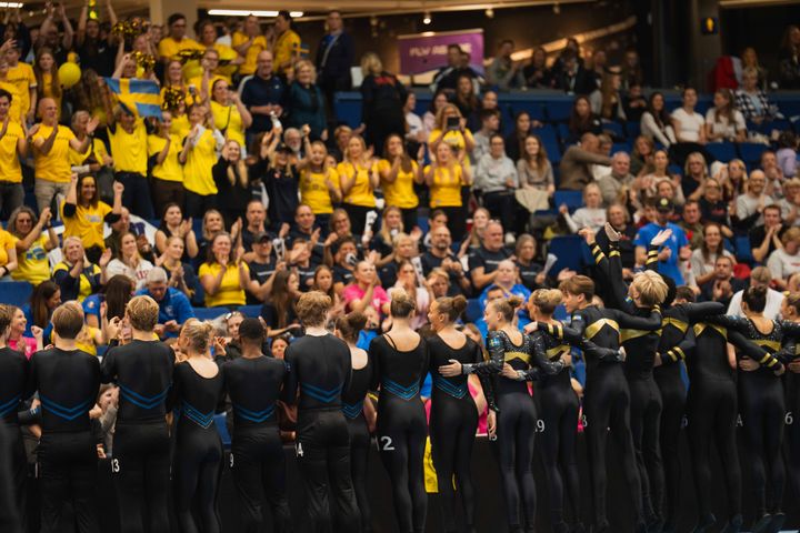 En grupp idrottare står i en arena och omfamnar varandra medan en publik klädd i gult och blått jublar i bakgrunden.