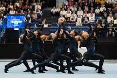 En grupp gymnaster i svarta dräkter utför en synkroniserad rörelse på en gymnastikmatta.
