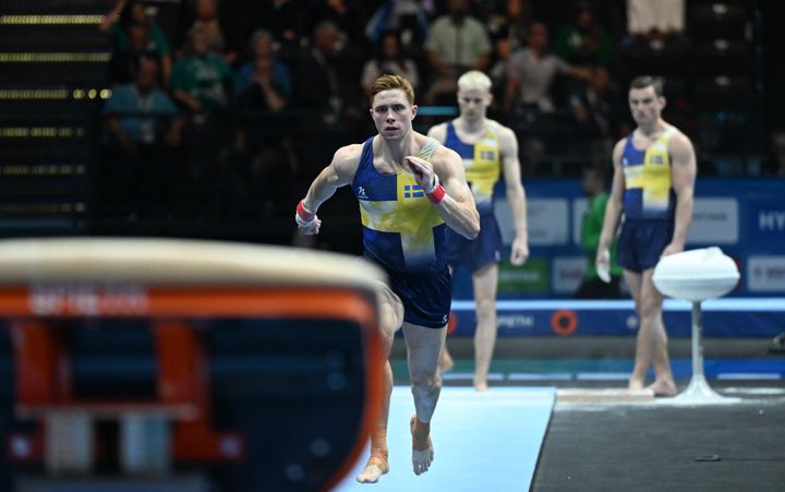 Gymnast springer mot hoppbordet under en tävling, med flera lagkamrater i bakgrunden.