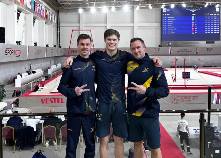 Tre glada män står tillsammans vid en gymnastikarena.