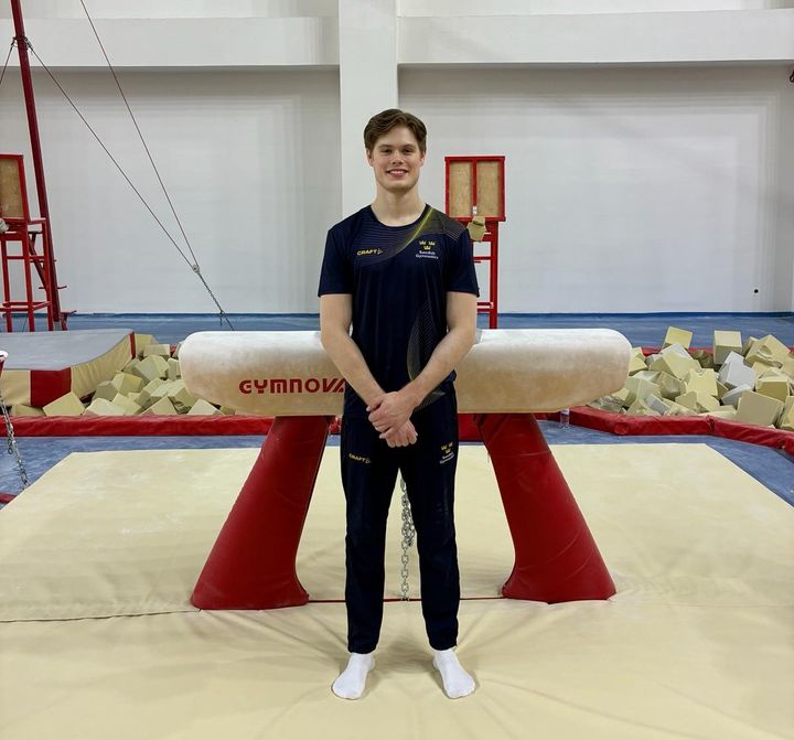 En man i träningskläder står leende framför en gymnastikbygel i en träningslokal.