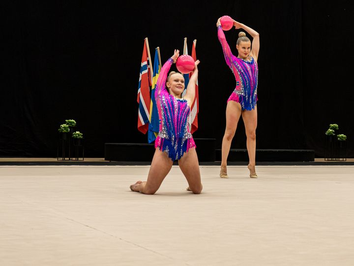 Två gymnaster i färgglada dräkter uppträder på en matta med bollar.