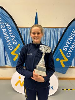 En kvinna håller en pokal framför en blå bakgrund med texten "Svensk Gymnastik".