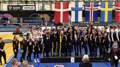 Grupp av unga gymnaster i svarta dräkter med gulddetaljer firar på prispallen med silvermedaljer och flaggor i bakgrunden.