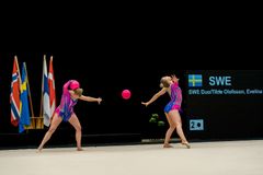 Två gymnaster i färgglada dräkter utför en rytmisk gymnastikövning med en boll på en tävlingsmatta, med flaggor och en tavla i bakgrunden.