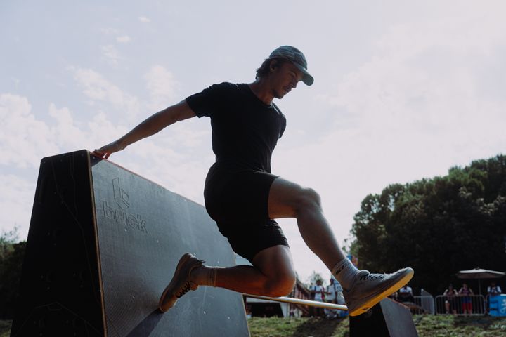 En person i mörka kläder och keps springer över ett hinder utomhus under en parkourtävling.