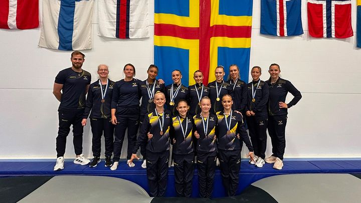Gruppfoto av det svenska kvinnliga gymnastiklaget som nyligen vann NM-guld, poserar framför nationella flaggor med sina medaljer.