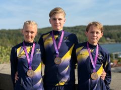 Tre ungdomar i likadana blå och gula sportjackor med medaljer runt halsen står framför en naturskön bakgrund med träd och vatten.