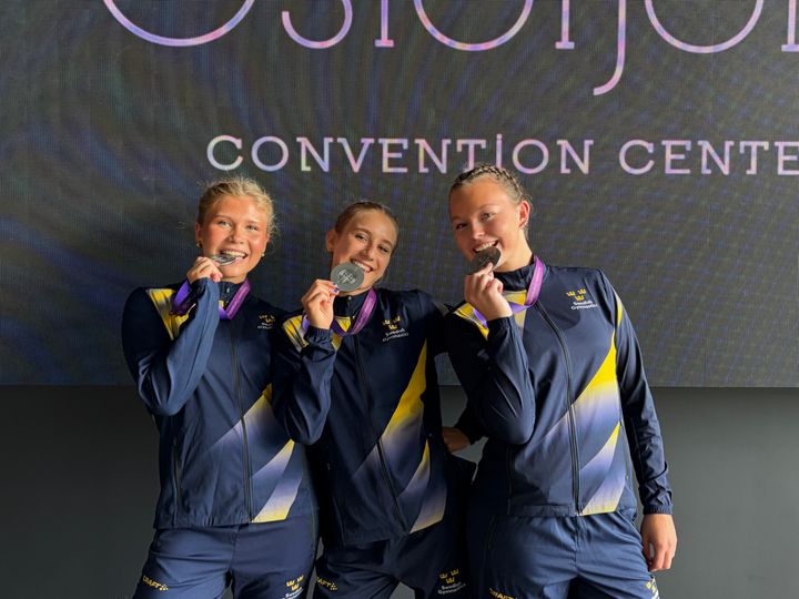 Tre personer i träningskläder poserar med medaljer vid ett evenemang.