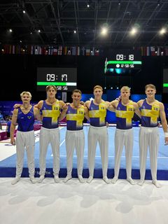 Sex manliga gymnaster från det svenska laget står på en gymnastikmatta i en arena, iklädda blå och gula tävlingskläder.