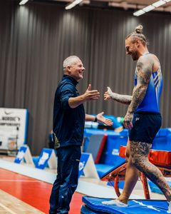 En tränare hälsar på en tatuerad idrottare i blå tävlingsdräkt vid en gymnastikarena.