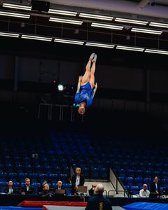 Gymnast i blå dräkt gör en volt högt över tävlingsmattan.