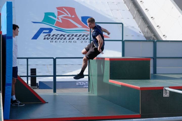 En parkourutövare hoppar mellan hinder på en tävlingsbana, med en skylt för "Parkour World Cup Beijing" i bakgrunden.