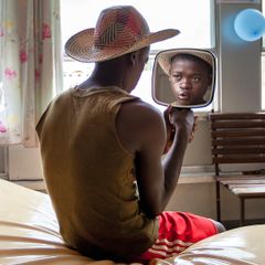 Tonårspojke i stråhatt sitter på en säng och ser sig i en liten spegel. Han har ett färskt ärr på överläppen.