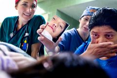 Flicka med färskt operationsärr på läppen ligger ner och bara hennes ansikte syns i liten spegel. Kvinna med tårar rinnande tittar på flickan. Bakom syns två sjukvårdspersonal.