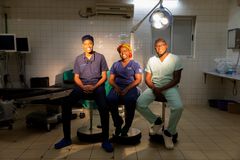 Tre kirurgklädda personer, två män och en kvinna, sitter på höga pallar i en operationssal.