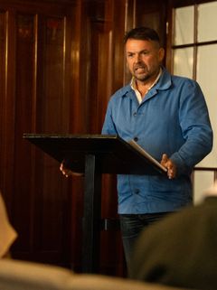 Man i blå skjorta står vid podium och ser ut att tala till en publik. Nästan helfigur.