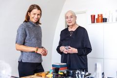 Kvinna klädd i grå stickad tröja och med en mörk halvlång page står bredvid en äldre kvinna klädd i svart, med grått hår i stram tofs. De tittar in i kameran och ser glada ut. I förgrund ett bord med lite prylar. I bakgrund vitt skåp med några glasföremål uppepå.