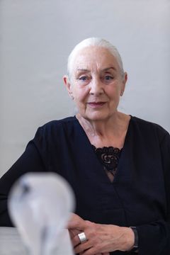 Kvinna klädd i svart, med grått hår i stram tofs, tittar in i kameran med ett lätt leende. I förgrund ett verk i glas.