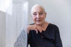 Kvinna klädd i svart, med grått hår i stram tofs, tittar in i kameran med ett lätt leende. Vilar hakan lätt mot ena handryggen. I förgrund en hög cylindervas.