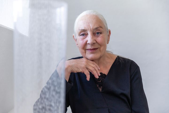 Kvinna klädd i svart, med grått hår i stram tofs, tittar in i kameran med ett lätt leende. Vilar hakan lätt mot ena handryggen. I förgrund en hög cylindervas.