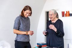 Kvinna klädd i grå stickad tröja och med en mörk halvlång page står bredvid en äldre kvinna klädd i svart, med grått hår i stram tofs. De tittar in i kameran och ser glada ut. I förgrund ett bord med lite prylar. I bakgrund vitt skåp med några glasföremål uppepå.