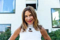 Kvinna med brunt axellångt hår ler mot kameran. I bakgrund skymtar ett vitt hus, av modernt snitt och med många fönster.