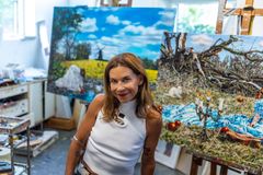 Kvinna med axellångt brunt hår och vit topp sitter i en målarateljé, framför en tavla på stafli.