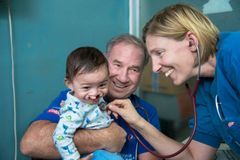 Blond kvinna i läkarkläder undersöker med sitt stetoskop ett litet barn, med mörk kalufs och läppspalt. Barnet sitter i en äldre mans knä. De ser glada ut.