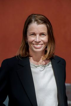 Mörkhårig kvinna, vit topp, svart kavaj, mörkröd fasad i bakgrund