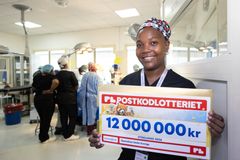 En leende kvinna iklädd scrubs står i sjukhusmiljö och håller upp en check som visar att Operation Smile har fått 12 miljoner från Postkodlotteriet.