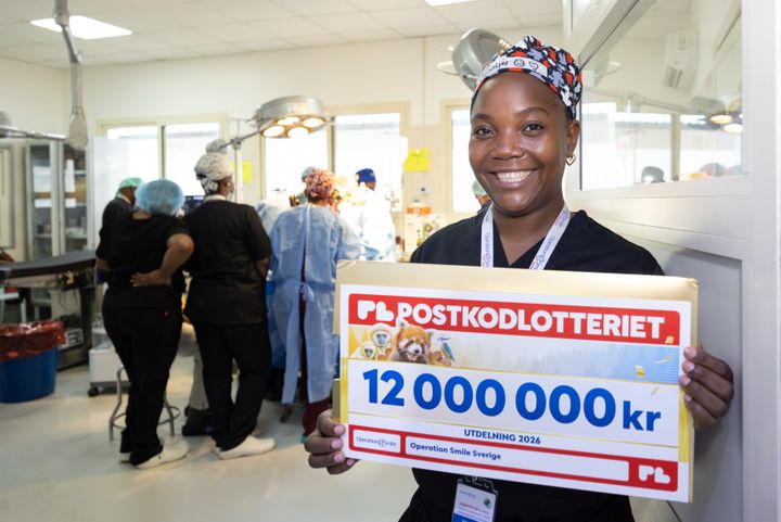 En leende kvinna iklädd scrubs står i sjukhusmiljö och håller upp en check som visar att Operation Smile har fått 12 miljoner från Postkodlotteriet.