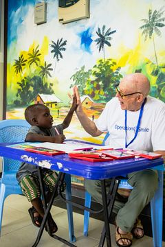 Vit man med glasögon och en Operation Smile-t-shirt gör high five med en ghanansk pojke. De sitter vid ett litet bord med ritsaker.