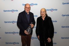 En man med en glaspjäs står bredvid en äldre kvinna framför en fotovägg med logotypen "Operation Smile".
