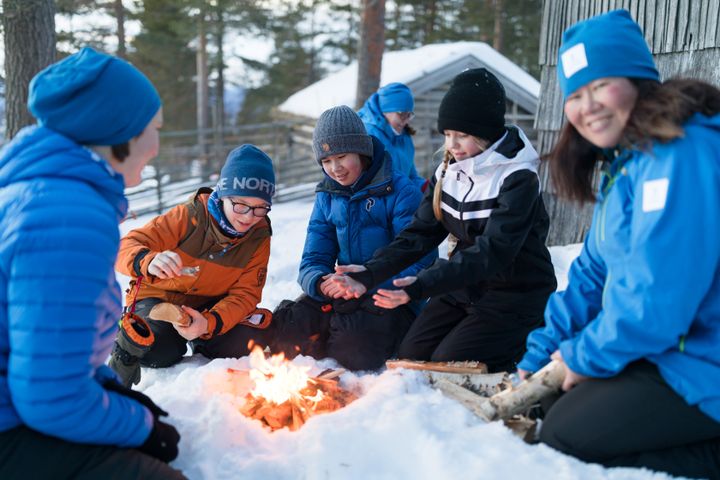 Barn och vuxna som värmer sig runt en eld i snöig natur.