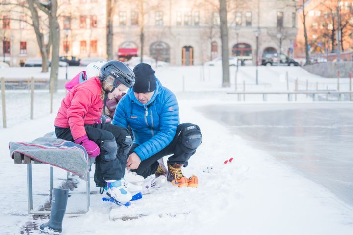 Ett barn som sitter på en bänk får hjälp av en vuxen man att sätta på sig skridskor.