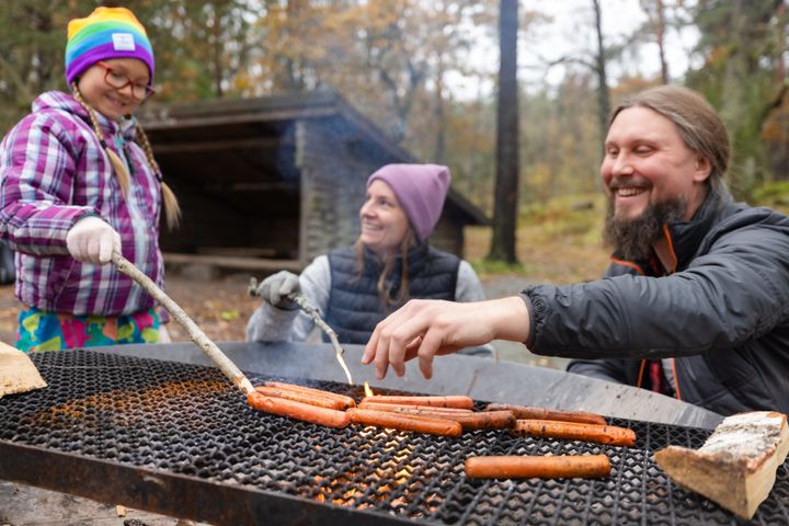 En barn och två vuxna som grillar korv vid en eldstad i ett naturområde.