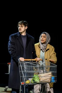 Två personer står vid en shoppingvagn på scenen, klädda i vinterkläder.