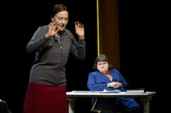 Två personer uppträder på en scen. En står i förgrunden medan den andra sitter vid ett bord i bakgrunden.