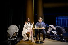 Två personer sitter vid ett bord på en scen, med barnvagnar på vardera sida.