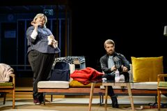 Två personer på en scen, en står och en sitter på en soffa, omgivna av rekvisita.